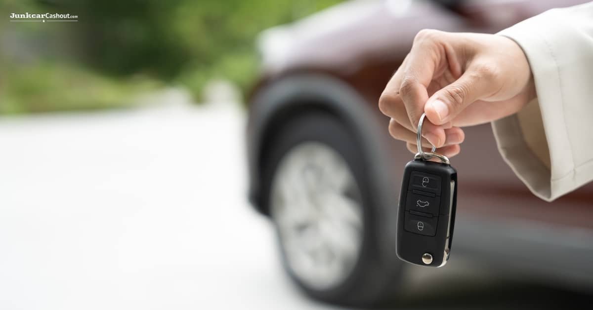 A person holds a car key fob in their hand with a brown vehicle parked in the background, considering where to sell car to get the most money.