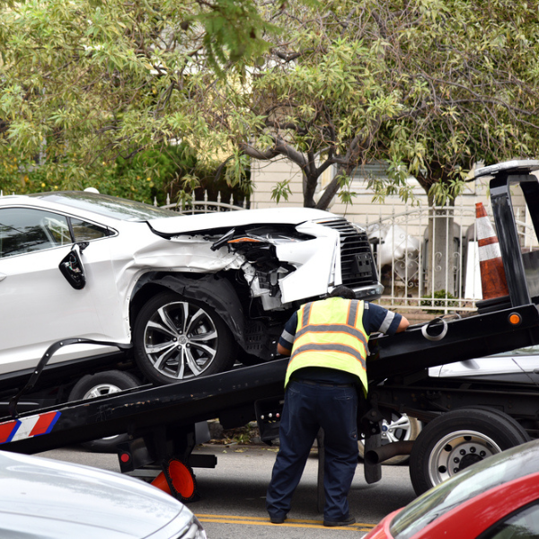 A tow truck operator loads a damaged white car with a crumpled front end onto a flatbed tow truck—just another day helping someone sell their junk car in Utah on a quiet residential street.